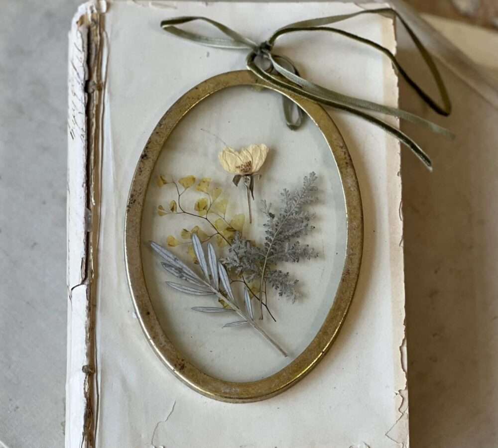 Gold Oval Pressed Flower Wall Art resting on a vintage book with a velvet ribbon hanger and delicate pressed botanicals inside glass.