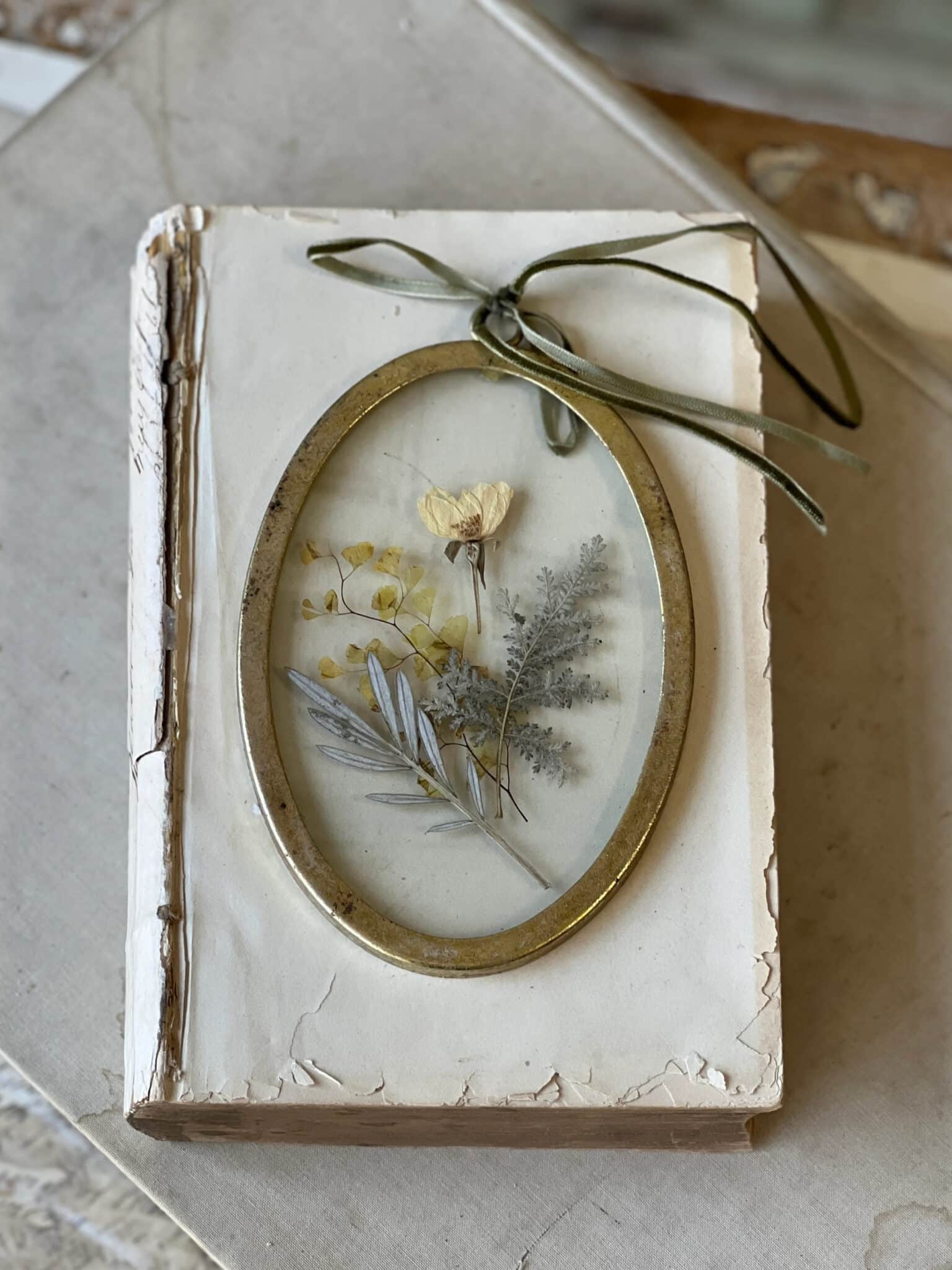 Gold Oval Pressed Flower Wall Art resting on a vintage book with a velvet ribbon hanger and delicate pressed botanicals inside glass.