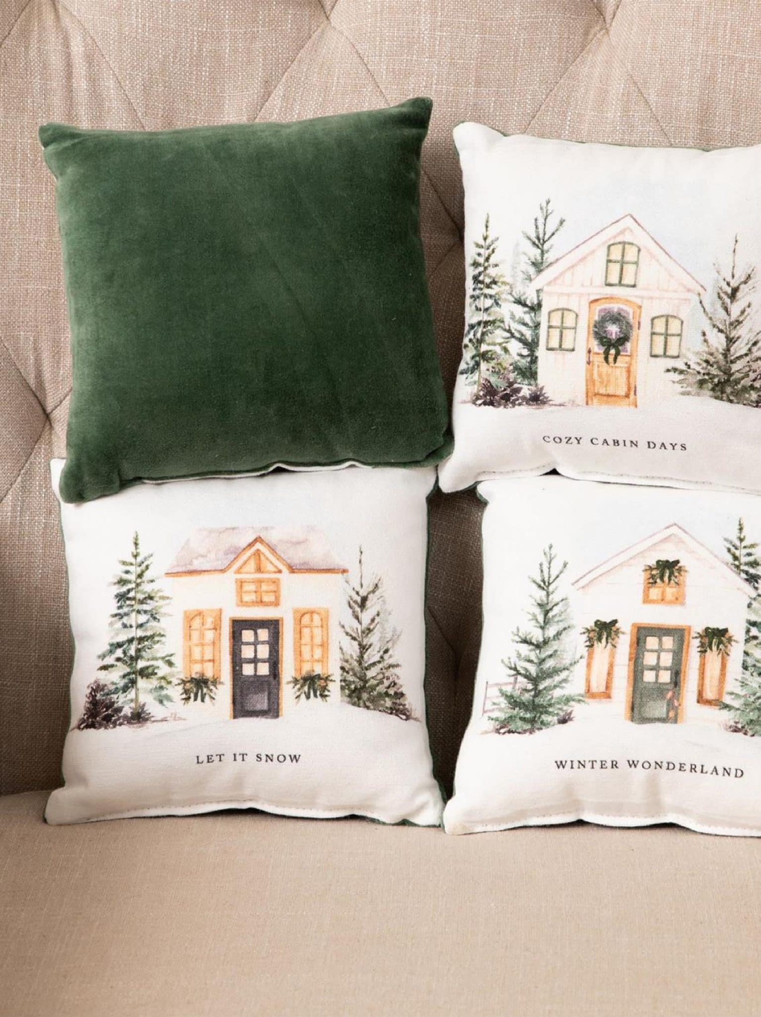 Set of small winter-themed pillows with snowy cottage illustrations and phrases “Cozy Cabin Days,” “Let It Snow,” and “Winter Wonderland,” shown with a green velvet pillow on a beige tufted sofa.