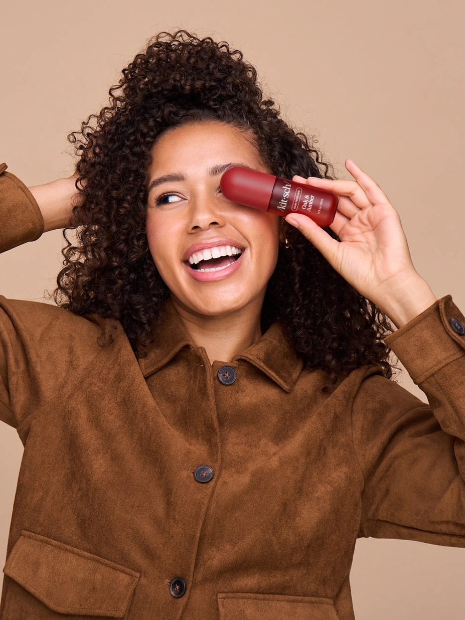 Woman smiling while holding Kitsch Oak & Amber Hair Perfume bottle near her face.