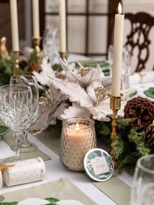 Shimmering Snowberry Candle glowing on holiday table with florals and pinecones.