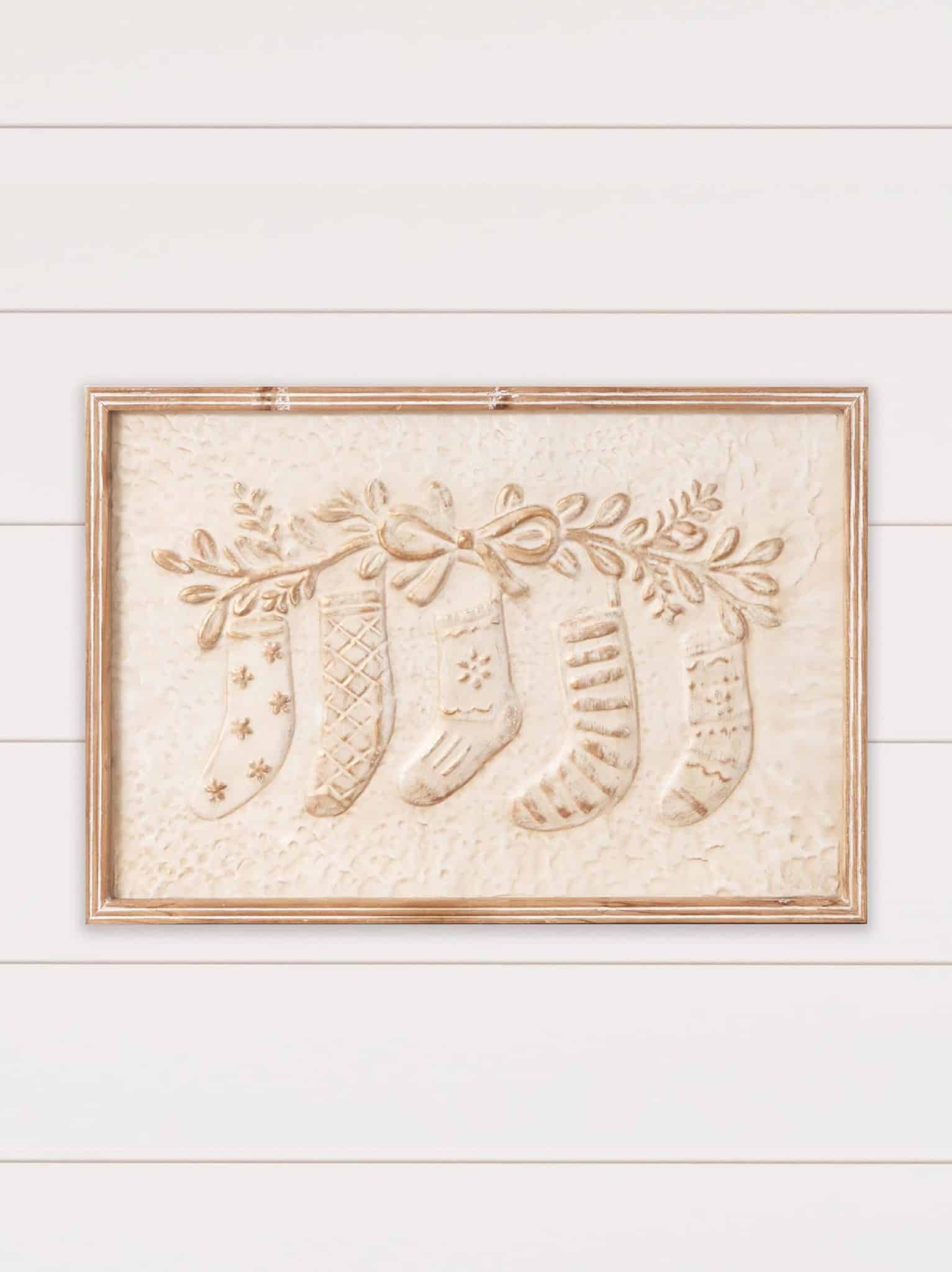 Embossed metal wall sign featuring raised Christmas stockings tied with bows and greenery, framed in natural wood and hung on a white shiplap wall.