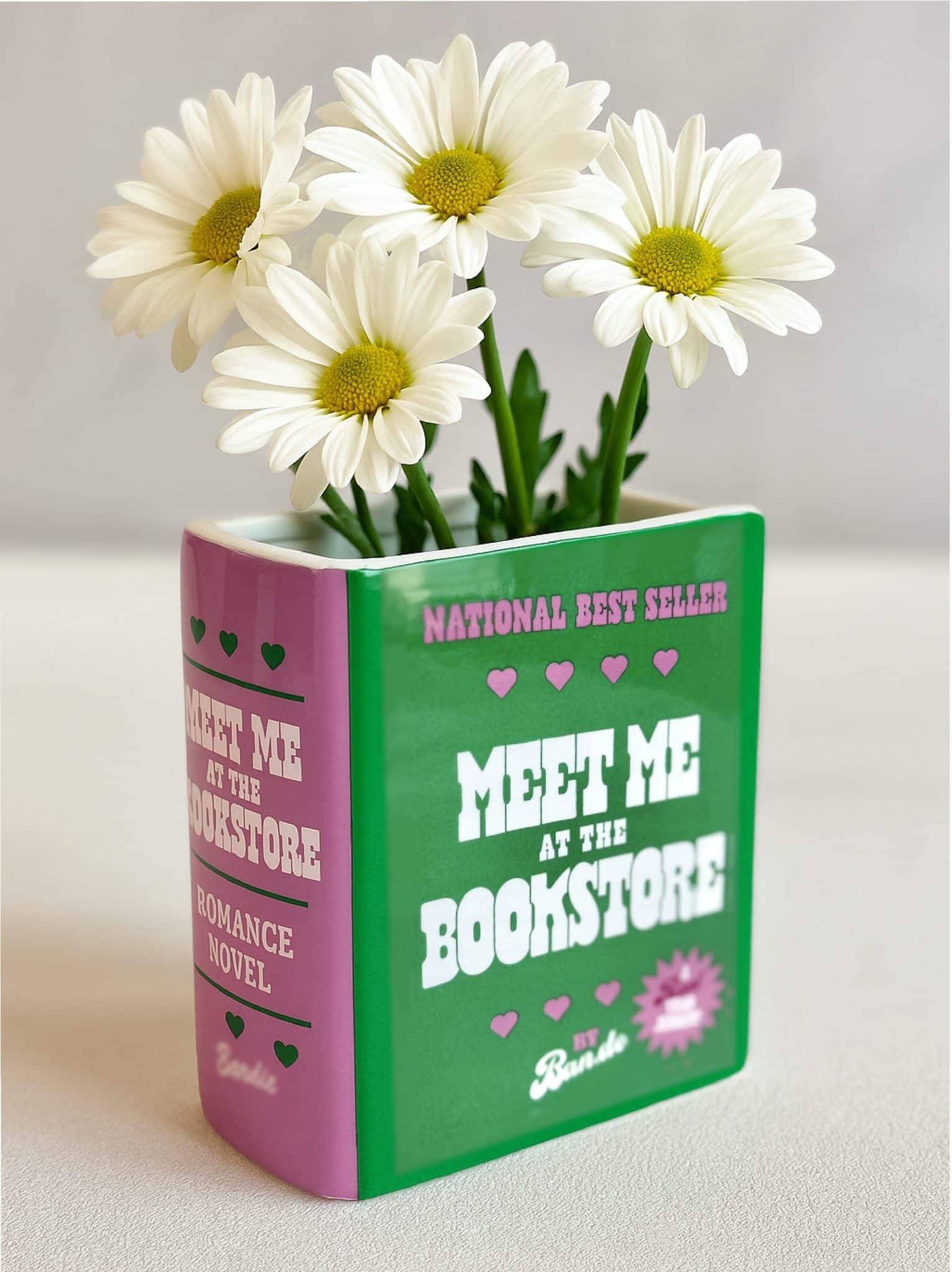 Ceramic book-shaped vase with the phrase "Meet Me at the Bookstore" filled with fresh white daisies.