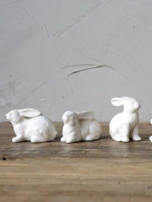 Three glossy white ceramic bunny figurines sitting in different poses on rustic wood tabletop.