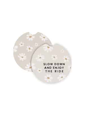 Round ceramic car coasters with white daisy designs and the phrase “Slow Down and Enjoy the Ride” on a soft beige background.