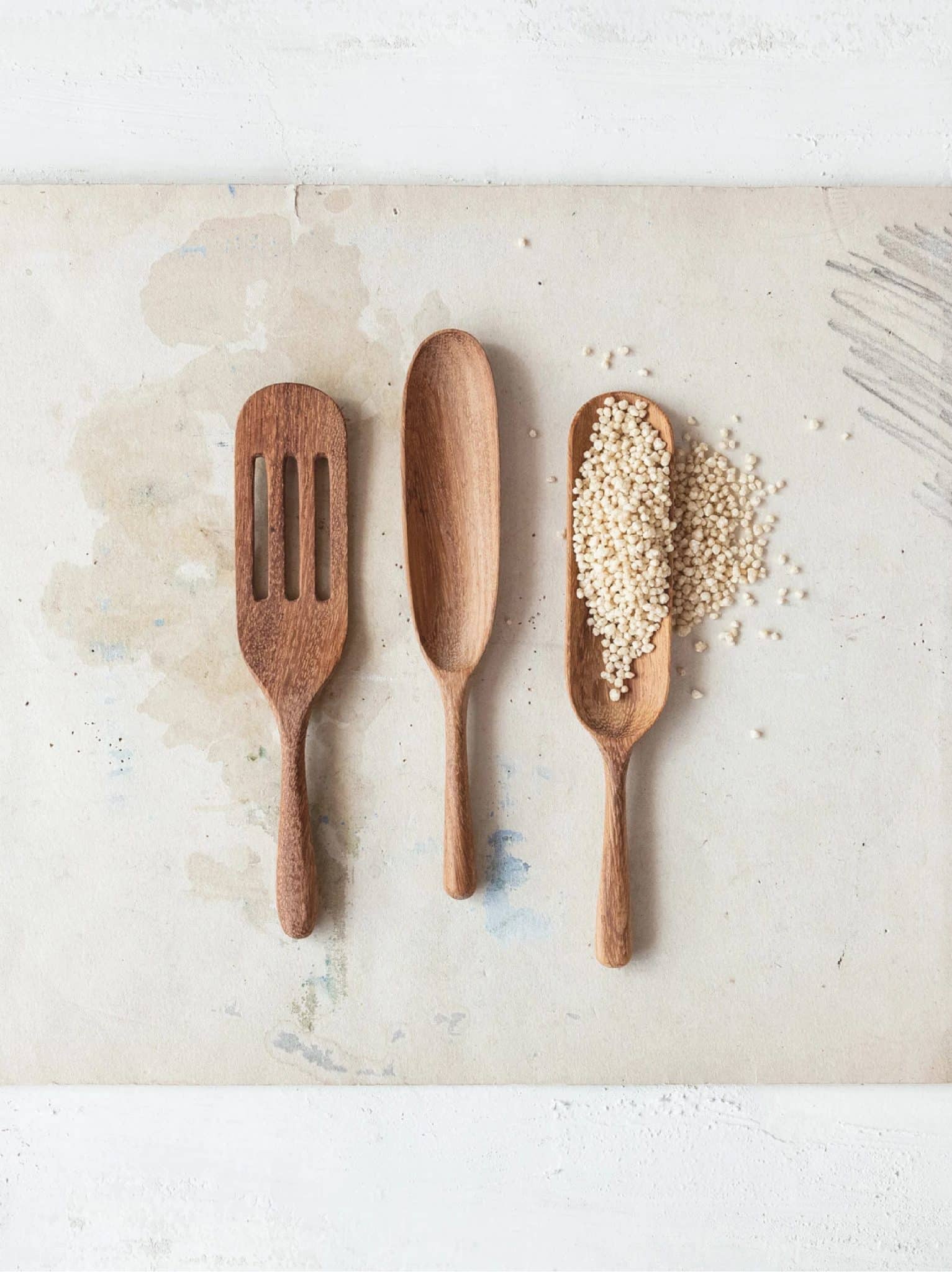 Three carved natural wood utensils including a spoon, spatula, and scoop displayed on a light surface with grains scattered for texture.
