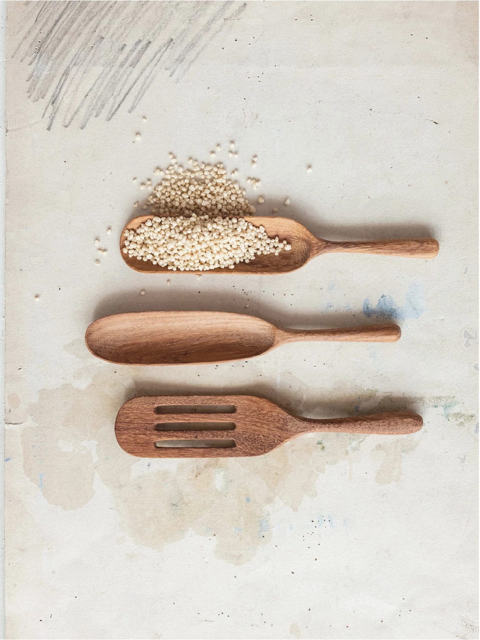 Three carved natural wood utensils, including a slotted spatula, displayed on a light surface with grains scattered for styling.