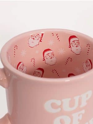 Interior of a light pink ceramic mug featuring red Santa faces and candy cane pattern inside, with “Cup of Cheer” text visible on the outside.