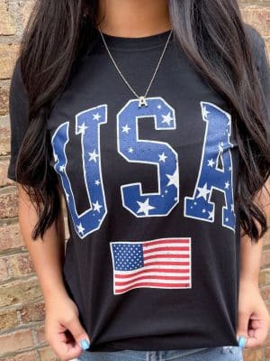 Close-up of a woman wearing a black graphic tee with bold “USA” star-filled lettering and a distressed American flag below, standing against a brick wall.