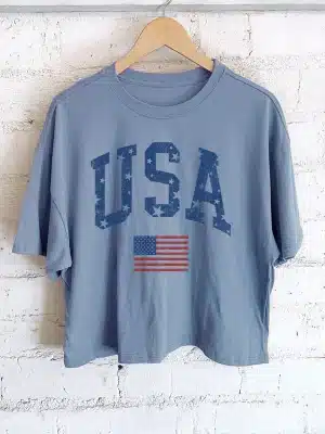 Indigo blue USA flag graphic long crop tee hanging on wooden hanger against white brick wall.