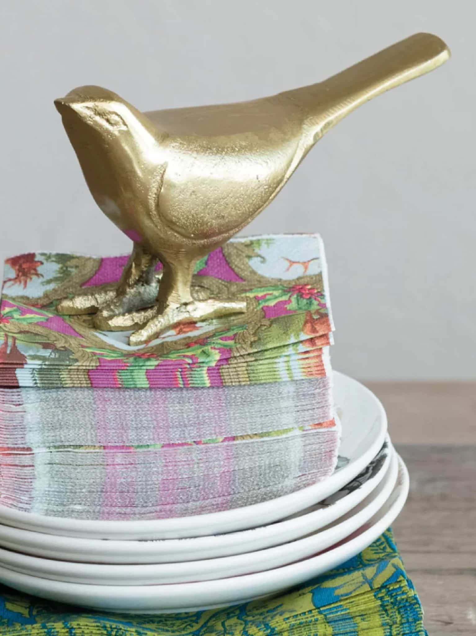 Smooth gold bird figurine sitting atop a stack of colorful napkins on a plate stack for decorative tabletop styling.