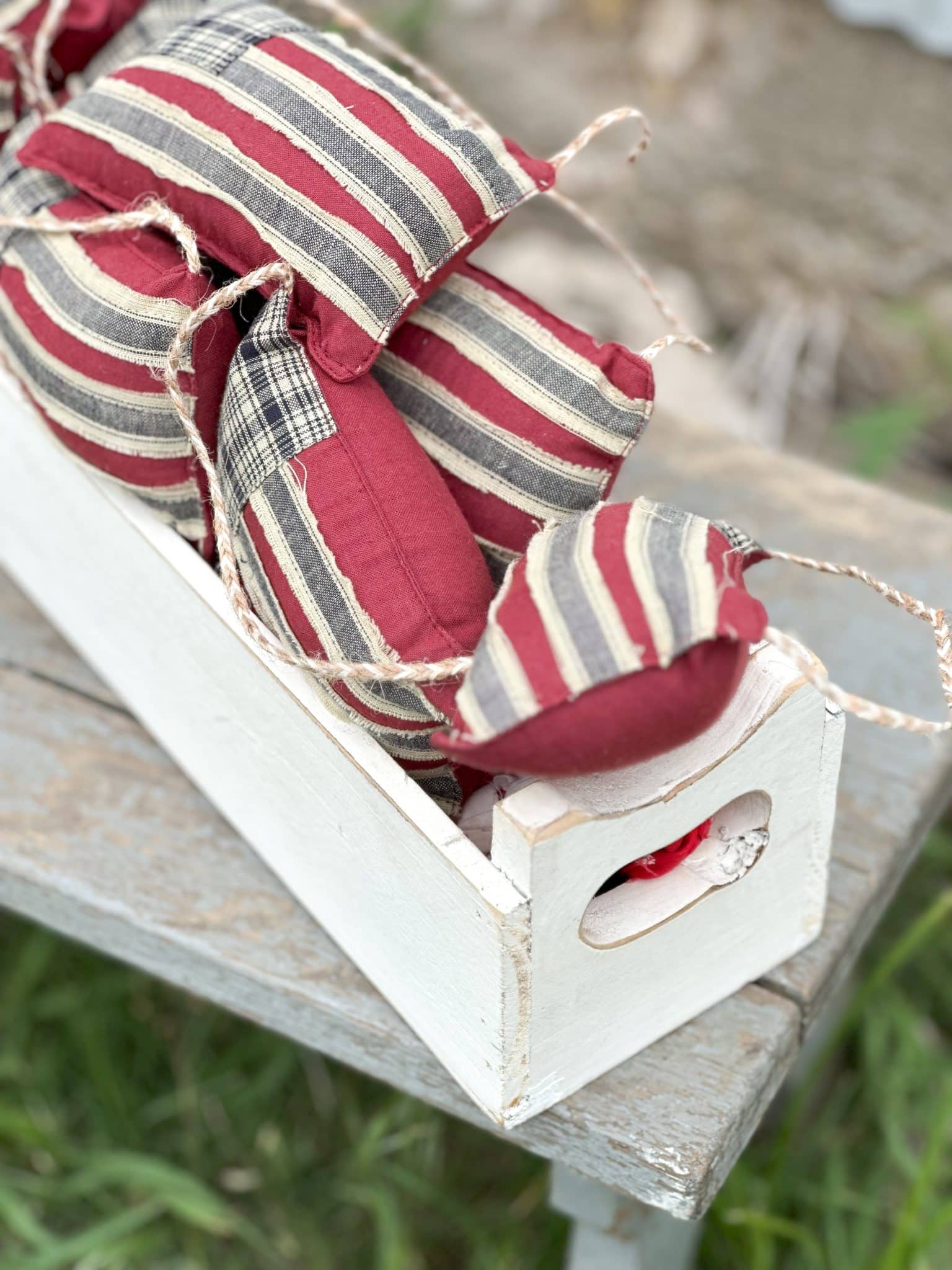Close-up of Mini Flag Bowl Fillers with red and plaid fabric inside a distressed white wooden tray on a rustic bench.