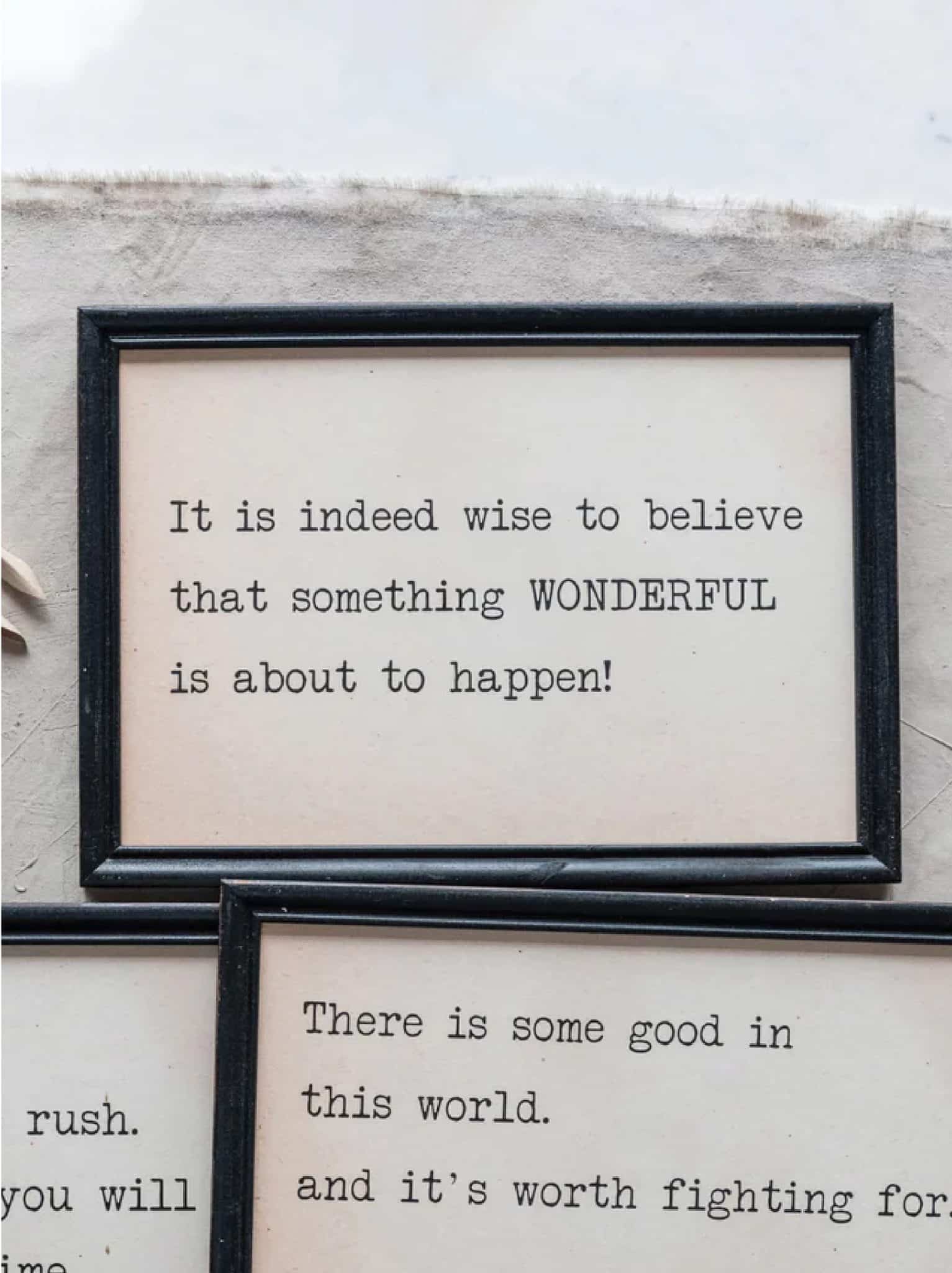 Decorative framed quote sign with cream background and black typewriter-style text reading “It is indeed wise to believe that something wonderful is about to happen!”