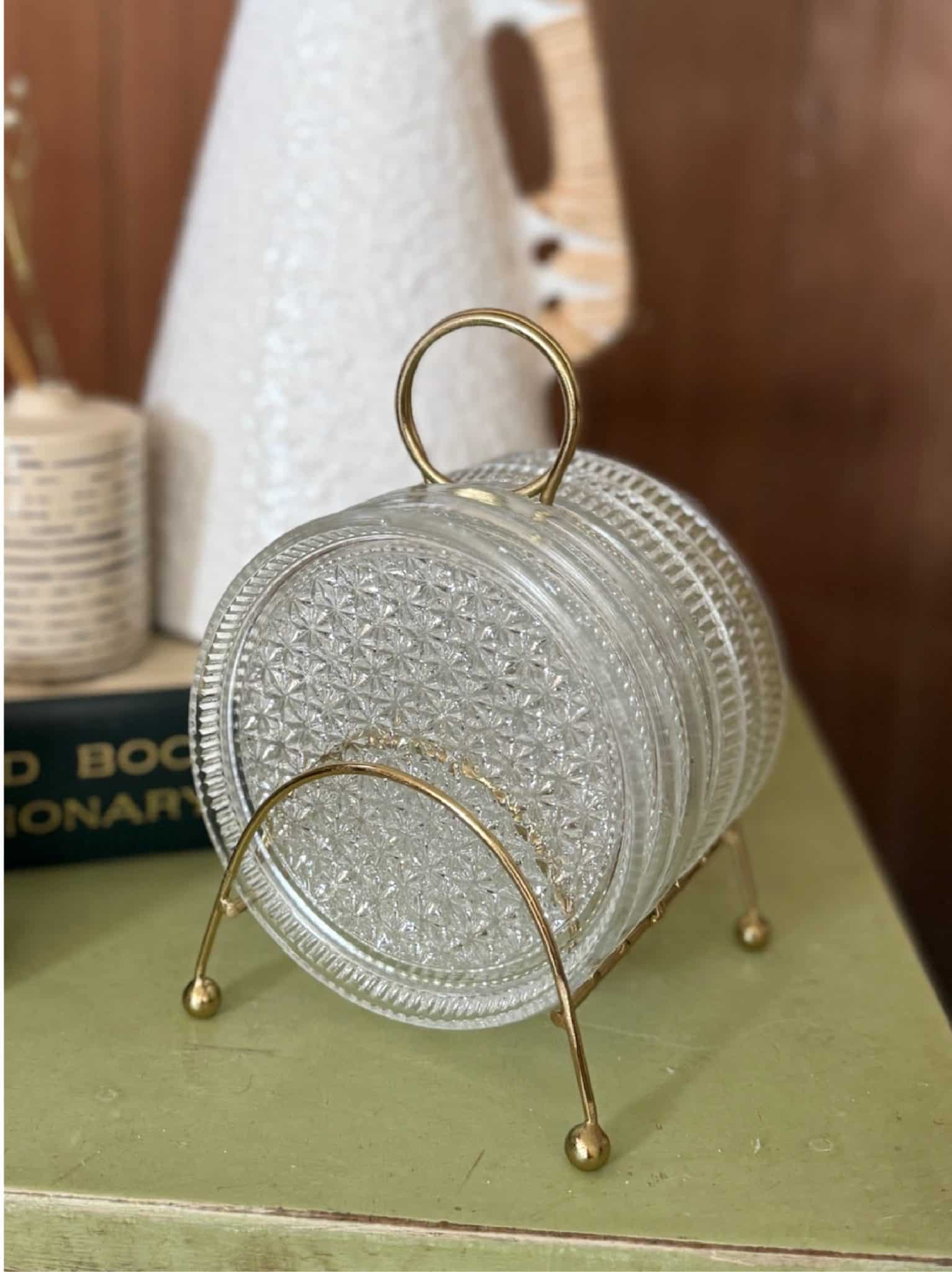 Close-up of clear pressed glass coasters with starburst pattern, held in a gold metal stand on a vintage green tabletop.