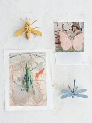 Four decorative pewter insect figurines—a bee, butterfly, grasshopper, and dragonfly—displayed on a white textured wall, each mounted over art prints and photographs for a playful, gallery-style presentation.