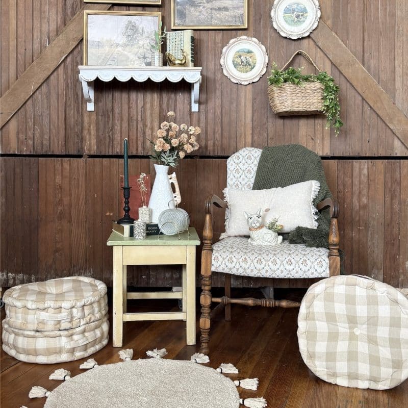Vintage wall gallery with gold and white frames, dog plates, hanging basket, rattan vase, bud vases, onion sprays, and cozy cottage accents including a tassel rug, buffalo check cushions, and a tufted armchair with green throw.