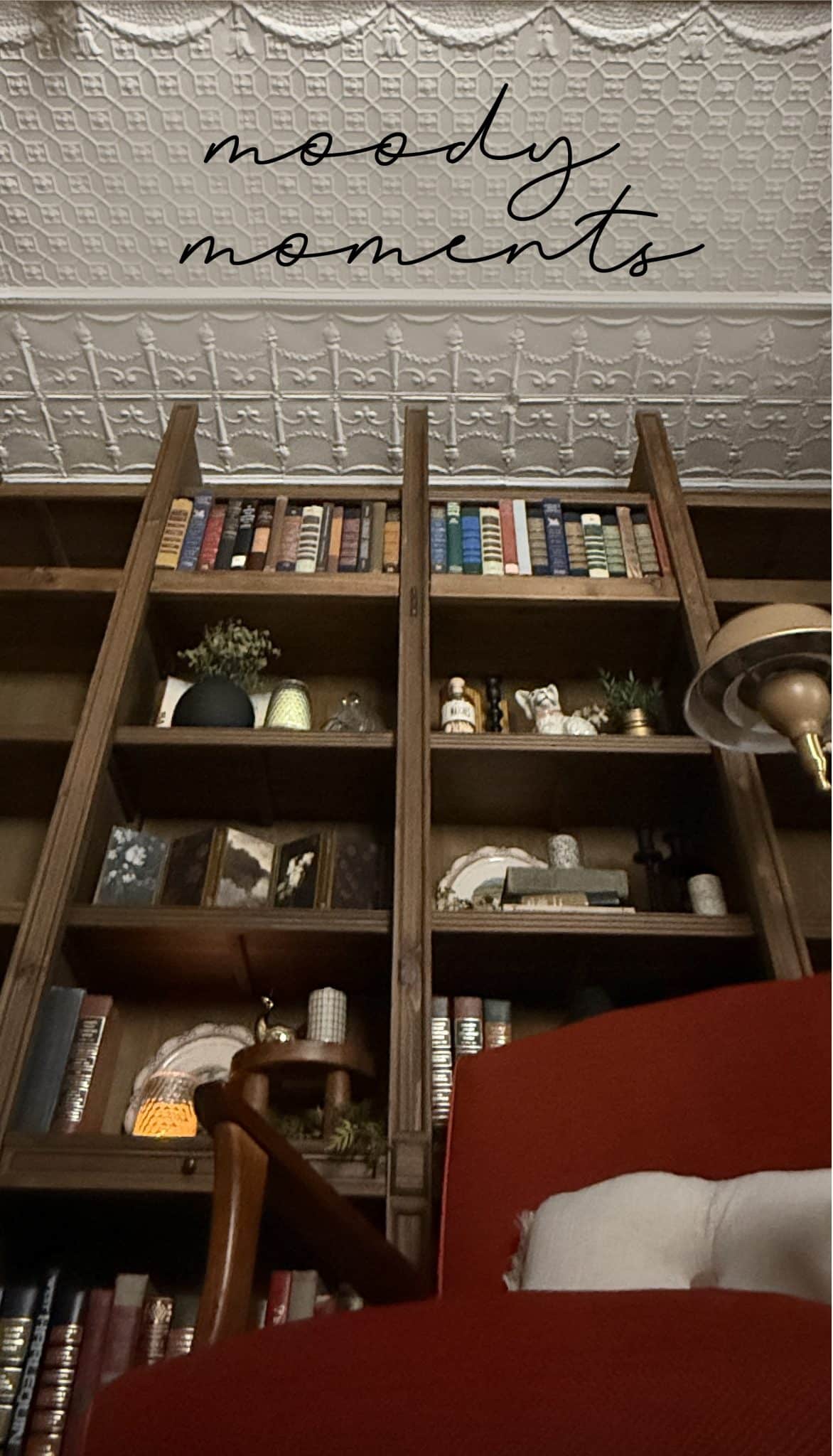 Tall wood bookshelf filled with vintage books, moody decor, and warm lighting under a white tin ceiling with the text “moody moments” overlayed at the top.