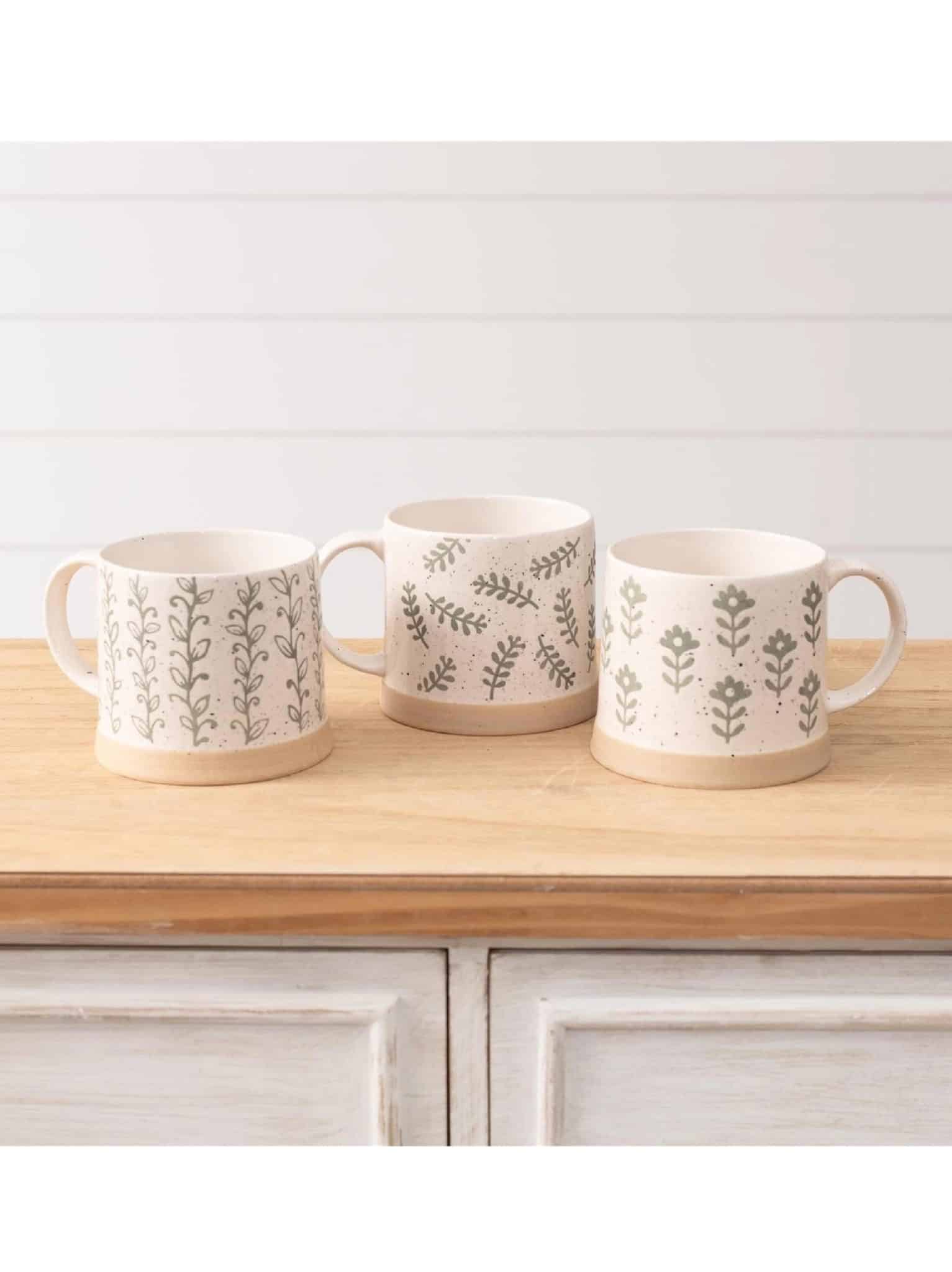 Hand-painted stoneware mugs with leafy green patterns in three styles displayed on a wood surface.