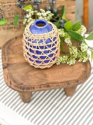 Indigo rattan-wrapped ceramic vase displayed on a rustic wood stool with white berry floral stems and greenery in the background.