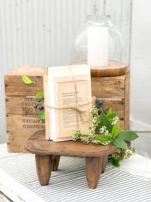 Vintage book bundle tied with twine on a small wood stool, styled with white berry florals and a glass hurricane candle holder in the background.
