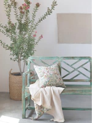 Cottage-style green wooden bench with floral throw blanket and pillow, cozy home decor setup with potted plant and neutral wall background.