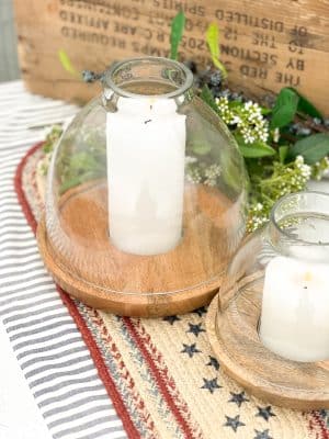 Large and small glass hurricane candle holders with wood bases on a patriotic jute table runner and ticking stripe runner