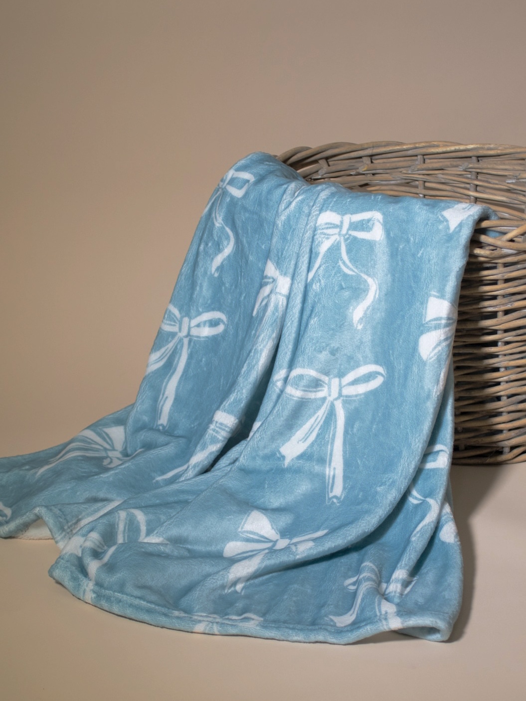 Light blue throw blanket with white bow pattern draped over a woven basket, showing its soft plush texture and elegant design.