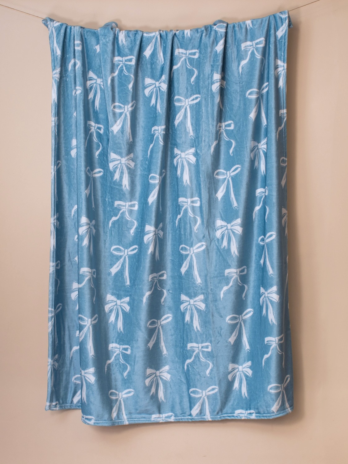 be-made-hays-ks-light-blue-bow-throw-blanket-full-view Light blue throw blanket with white bow pattern hanging to show full length and plush texture against a neutral background.