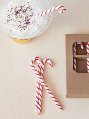 Red and white Glass Candy Cane Stir Sticks shown beside a gift box and a bowl of whipped cream for festive holiday drinks.