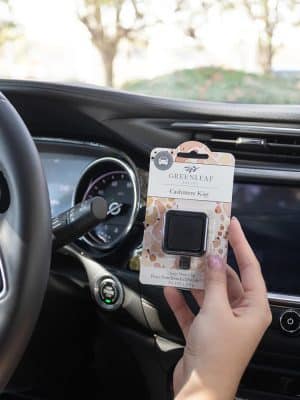Hand holding Cashmere Kiss Auto Vent Clip packaged inside car by steering wheel.