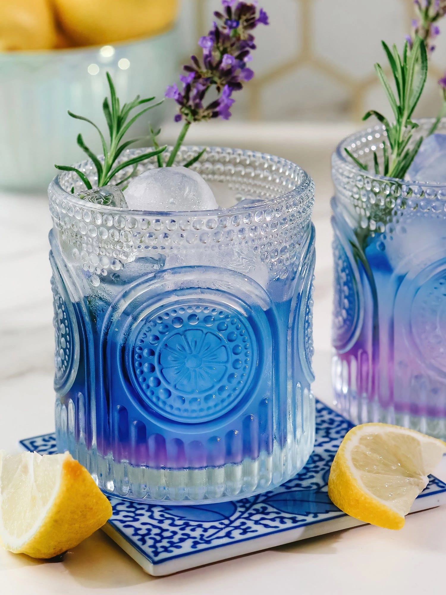 Clear retro arched drinking glass holding a vibrant blue cocktail with ice, rosemary, and lemon garnish.