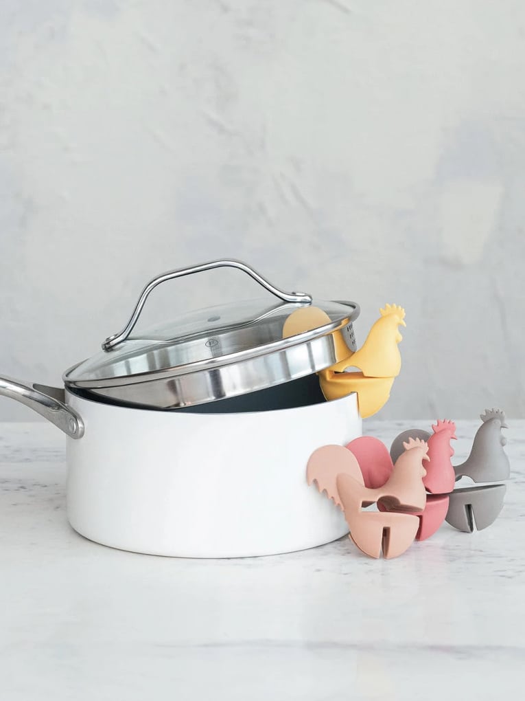 Close-up of yellow, tan, rose, and grey silicone rooster spoon holders on a white pot with metal lid.