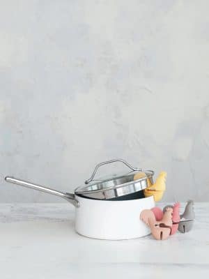 Assorted silicone rooster spoon holders in yellow, tan, rose, and grey shown on a white pot with lid in a neutral kitchen setting.