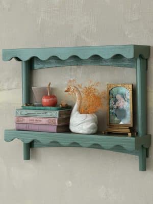 Teal wood scalloped wall shelf styled with books, framed art, a small vase, and a decorative swan figurine against a neutral wall.