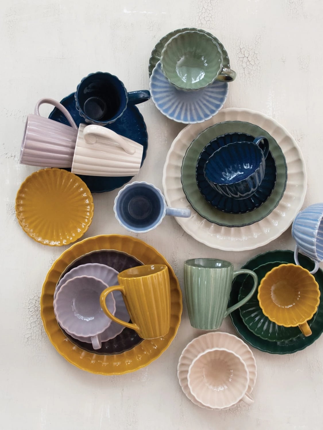 Assorted stoneware ribbed mugs in cream, sage green, and light blue displayed among matching bowls and plates on a neutral background.