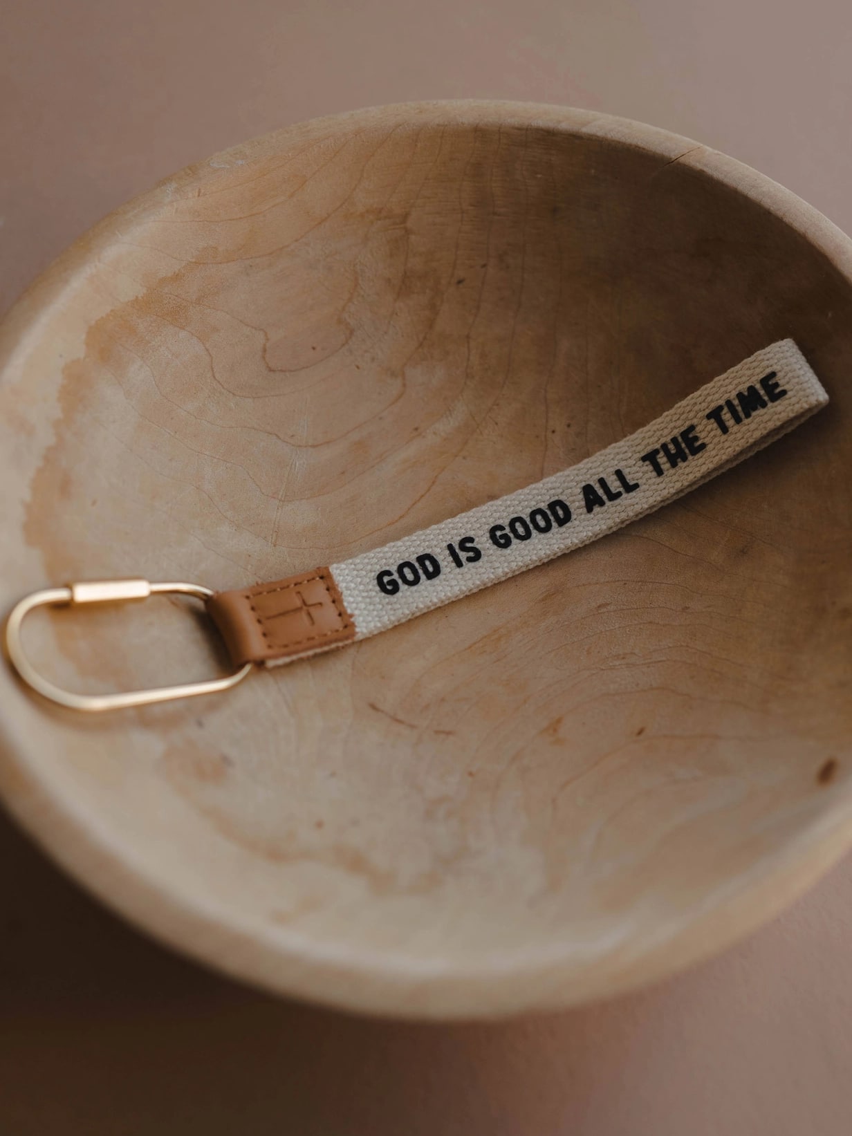 God is Good All the Time wristlet keychain resting in a wood bowl with gold carabiner and cross-stamped leather tag.