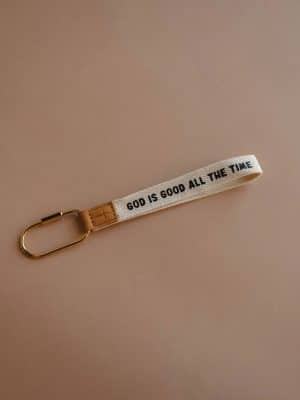 Neutral background photo of the God is Good All the Time keychain wristlet with woven strap, leather tag, and gold carabiner.