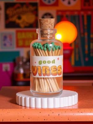 Glass jar of green-tipped matches labeled “Good Vibes” in retro lettering, topped with a cork and styled against a colorful, vintage-inspired backdrop.