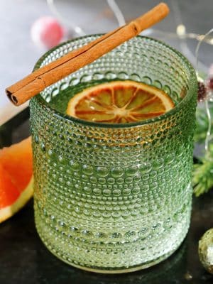 Sage green beaded old fashion glass with cocktail, dried orange slice, and cinnamon stick garnish.