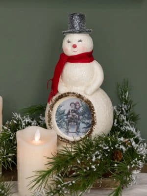 Vintage-inspired snowman figurine with silver top hat, red scarf, and glitter finish, featuring a tinsel-framed winter print at the center.