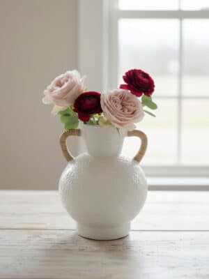 A medium ceramic vase with rattan-wrapped handles holding red and blush flowers on a light wood table near a window.