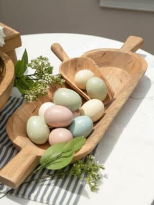 A carved wooden dough bowl with large handles holds pastel eggs with fresh greenery for a spring centerpiece on a table.