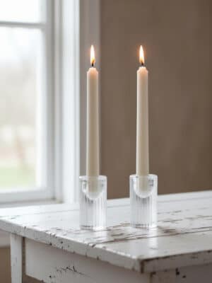A pair of clear cut-glass taper candle holders with lit ivory taper candles styled on a white table by a window.