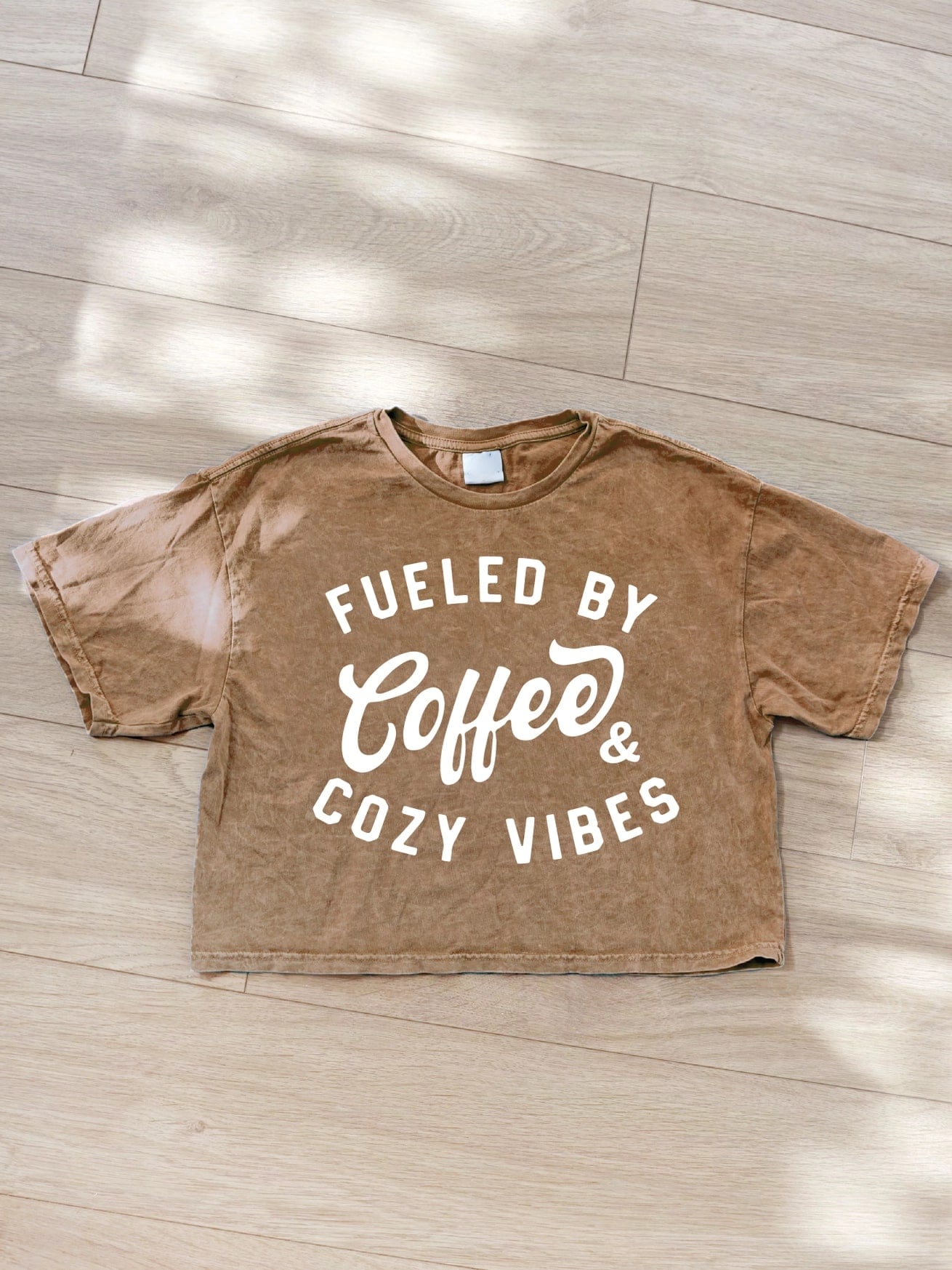 Toast tan cropped graphic tee with white lettering that reads “Fueled by Coffee & Cozy Vibes,” mineral washed cotton jersey.
