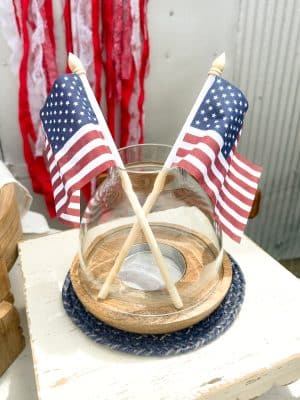 Glass hurricane candle holder with wooden base, styled with crossed American flag picks and placed on a blue braided jute trivet.