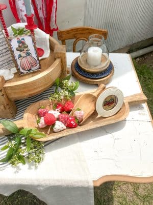 Patriotic summer vignette featuring a wood dough bowl filled with fabric strawberries, a scoop, a vintage July 4th tin, and a hurricane candle holder on a white chippy table.