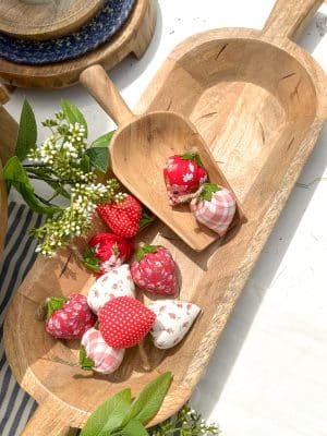 Carved wooden dough bowl filled with red and white fabric strawberries styled with a scoop.