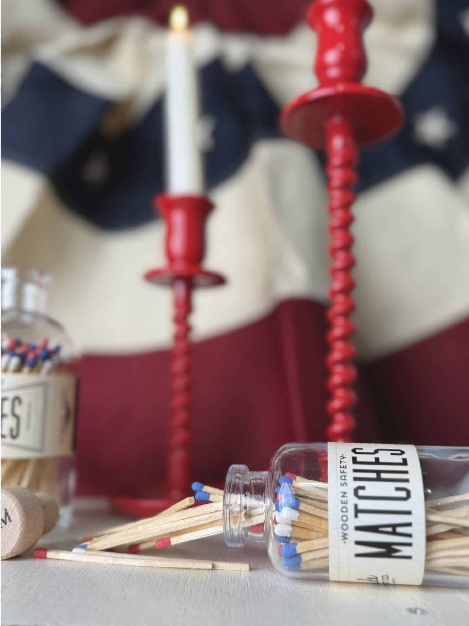 Mini and full-size USA match jars with red, white, and blue tips styled alongside red corkscrew taper candle holders and a lit white taper candle in a patriotic scene.