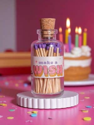Mini Apothecary glass jar of purple-tipped matches with a cork top and “Make a Wish” retro-style label, styled on a pink table with birthday candles and cake in the background.