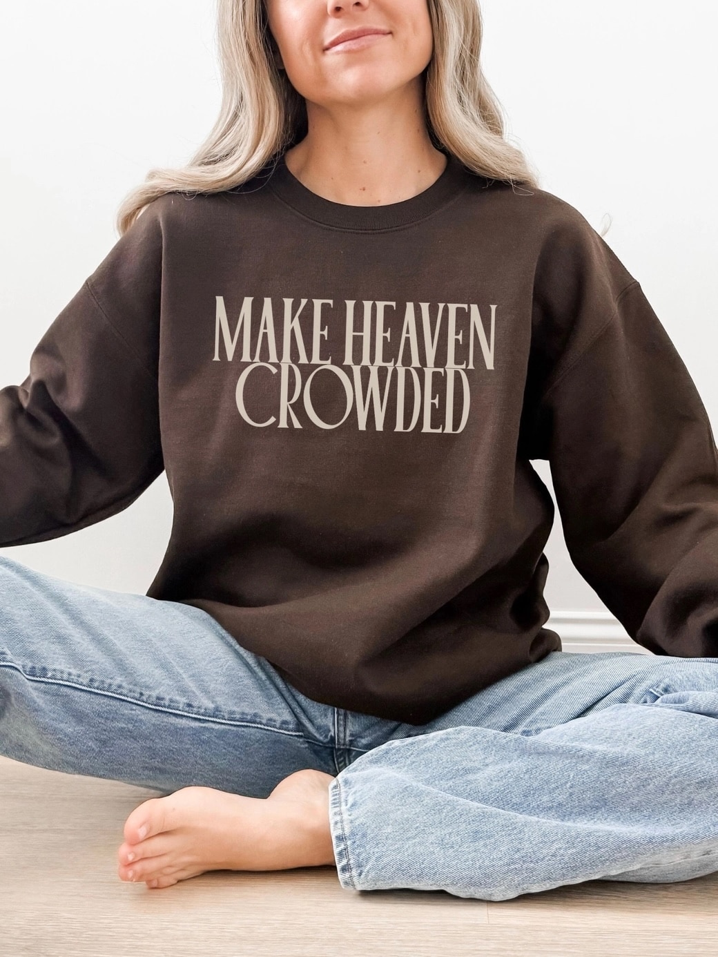 Woman wearing a chocolate brown “Make Heaven Crowded” crewneck sweatshirt with bold block lettering, styled casually with light denim jeans.